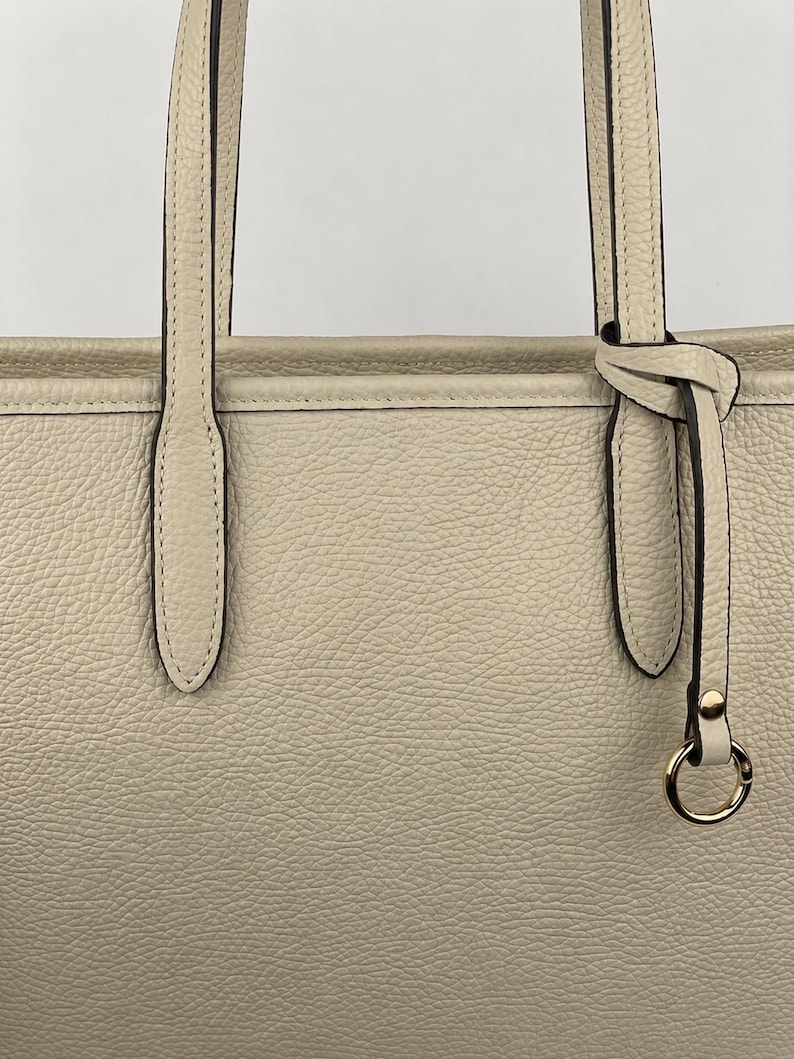 Op de afbeelding: Een beige tote bag met een generfde textuur, voorzien van twee verticale schouderbanden bevestigd aan de bovenrand. Een dun leren koord is rond &eacute;&eacute;n band geknoopt en eindigt in een kleine goudkleurige metalen ring met bijpassende klinknagel.