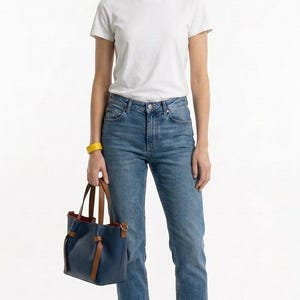 May include: A white crew neck t-shirt, blue denim jeans, white sneakers, a yellow wristband, and a navy blue handbag with brown leather handles and a red interior lining. The handbag features a knotted strap design on the front exterior.