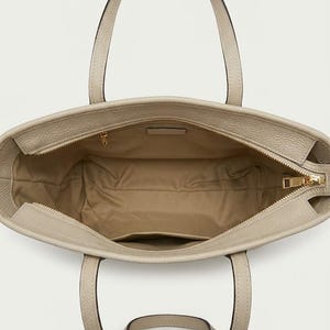 Op de afbeelding: Een beige tote bag met een textuur aan de buitenkant en twee handvatten. De binnenzijde is gevoerd met een lichtbruine stof en is voorzien van een ritsvak met een goudkleurige metalen trekker en een klein open steekvak aan de zijkant.