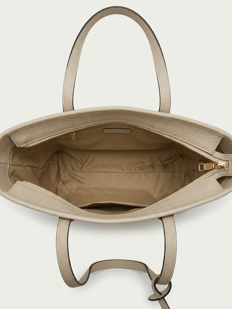 Op de afbeelding: Een beige tote bag met een textuur aan de buitenkant en twee handvatten. De binnenzijde is gevoerd met een lichtbruine stof en is voorzien van een ritsvak met een goudkleurige metalen trekker en een klein open steekvak aan de zijkant.