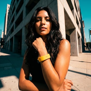 Peut inclure: Un bracelet en silicone jaune vif pr&eacute;sentant un petit d&eacute;tail d&eacute;coup&eacute; rectangulaire. Le bracelet est port&eacute; sur l'avant-bras et poss&egrave;de une finition mate et lisse, offrant une touche de couleur vive sur les v&ecirc;tements sombres de la personne.