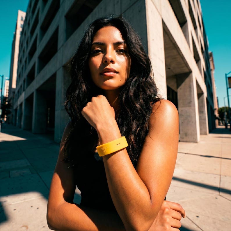 Peut inclure: Un bracelet en silicone jaune vif pr&eacute;sentant un petit d&eacute;tail d&eacute;coup&eacute; rectangulaire. Le bracelet est port&eacute; sur l'avant-bras et poss&egrave;de une finition mate et lisse, offrant une touche de couleur vive sur les v&ecirc;tements sombres de la personne.