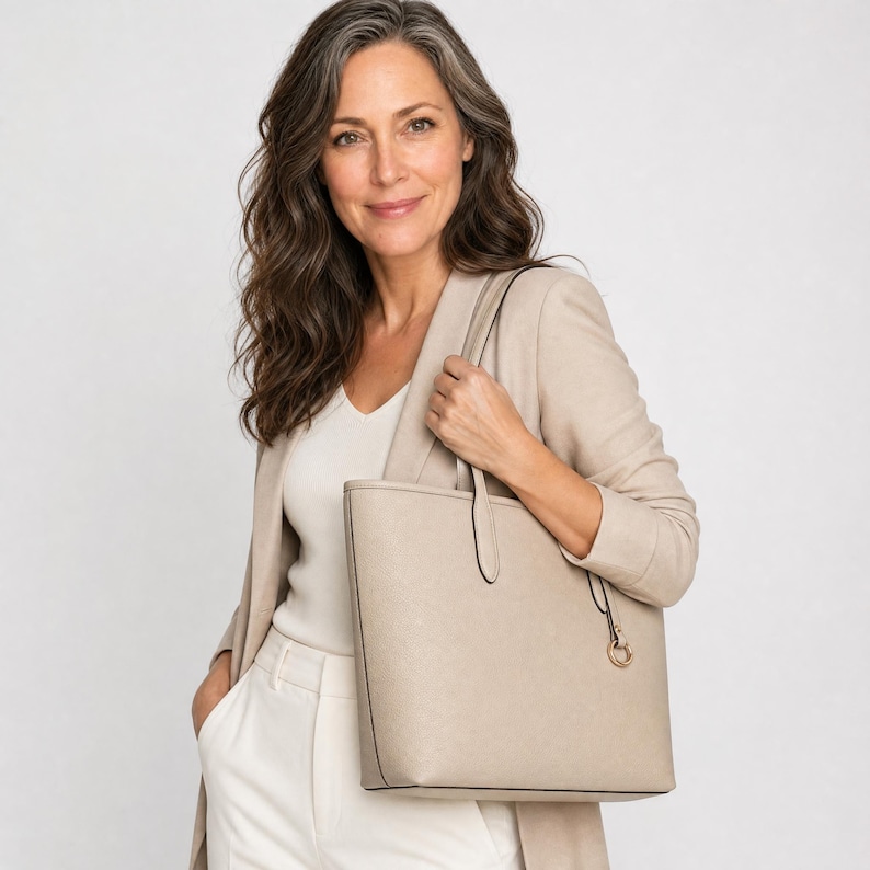 Op de afbeelding: Een gestructureerde beige tote bag met een gladde, matte afwerking, voorzien van twee dunne schouderbanden en een kleine, ronde goudkleurige metalen bedel die aan een van de bandbevestigingen hangt. De tas heeft een strak, rechthoekig silhouet.