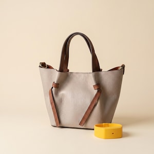 Peut inclure: Un sac &agrave; main en cuir gris clair dot&eacute; de deux anses sup&eacute;rieures marron fonc&eacute; et de liens lat&eacute;raux en cuir marron assortis qui resserrent les c&ocirc;t&eacute;s. Un bracelet de suivi d'activit&eacute; en silicone jaune vif est plac&eacute; devant le sac.