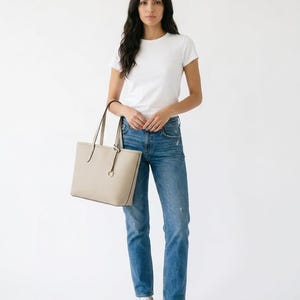 Op de afbeelding: Een beige gestructureerde tote bag met dunne schouderbanden en een ronde metalen bedel, gecombineerd met een effen wit T-shirt met korte mouwen, een blauwe spijkerbroek met een versleten afwerking en strakke witte sneakers.