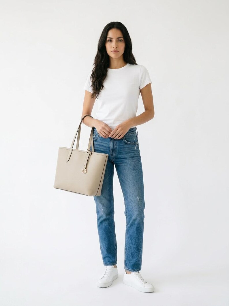 Op de afbeelding: Een beige gestructureerde tote bag met dunne schouderbanden en een ronde metalen bedel, gecombineerd met een effen wit T-shirt met korte mouwen, een blauwe spijkerbroek met een versleten afwerking en strakke witte sneakers.