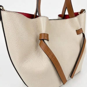 K&ouml;nnte beinhalten: Eine beigefarbene Handtasche mit strukturierter Oberfl&auml;che, braunen Ledergriffen und dekorativen, geknoteten Seitenelementen aus braunem Leder. Das Innere der Tasche ist mit einem leuchtend roten Material ausgekleidet, das oben sichtbar ist.