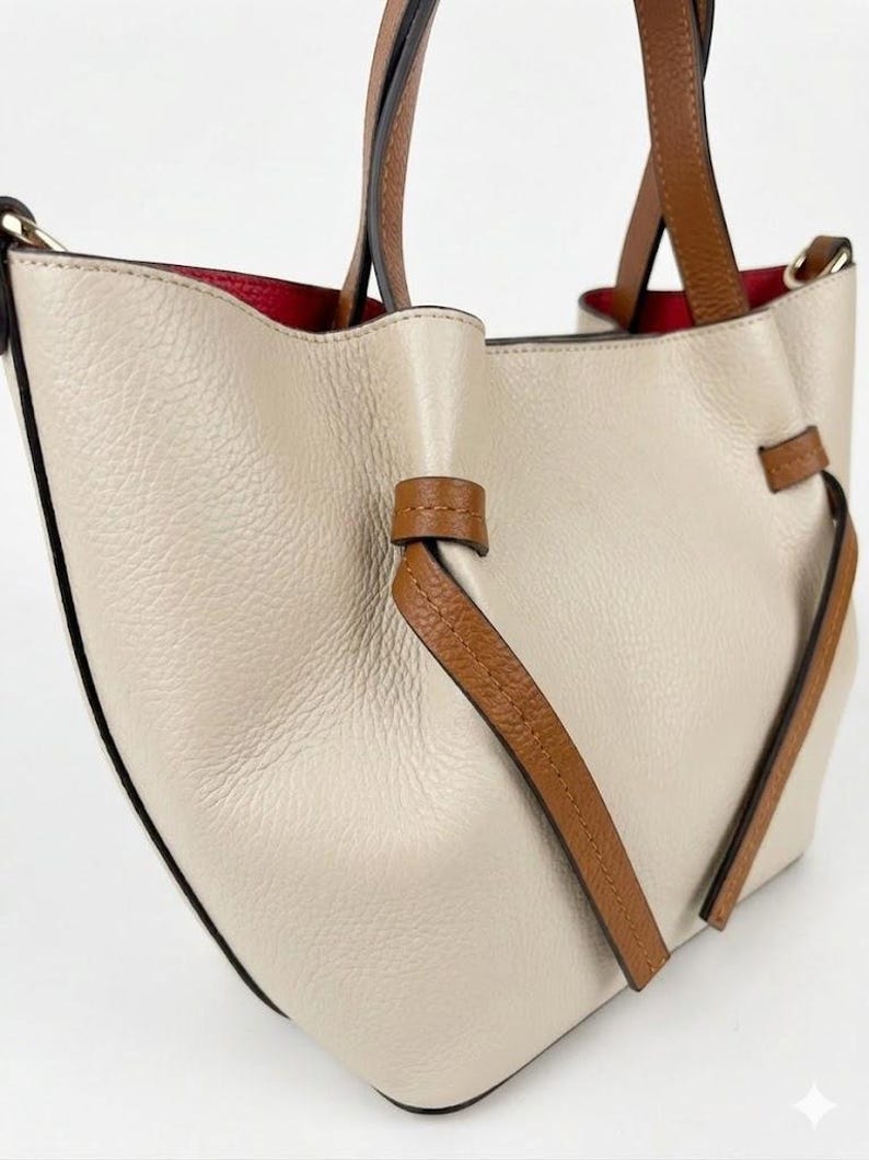 K&ouml;nnte beinhalten: Eine beigefarbene Handtasche mit strukturierter Oberfl&auml;che, braunen Ledergriffen und dekorativen, geknoteten Seitenelementen aus braunem Leder. Das Innere der Tasche ist mit einem leuchtend roten Material ausgekleidet, das oben sichtbar ist.