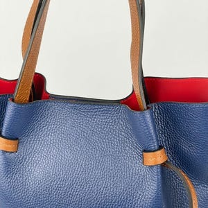 May include: A blue pebbled leather handbag featuring two tan leather handles and a vibrant red interior lining. The exterior includes a tan leather strap detail threaded through the sides of the bag, creating a cinched effect at the top opening.