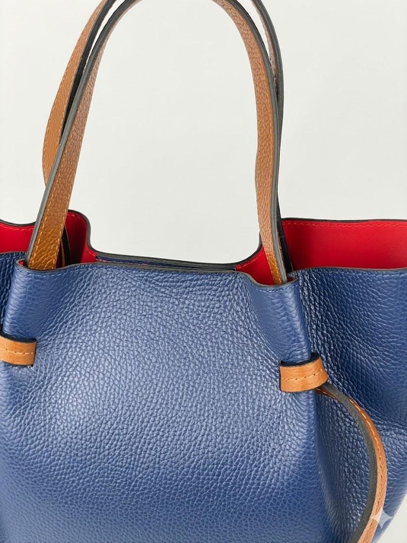 May include: A blue pebbled leather handbag featuring two tan leather handles and a vibrant red interior lining. The exterior includes a tan leather strap detail threaded through the sides of the bag, creating a cinched effect at the top opening.