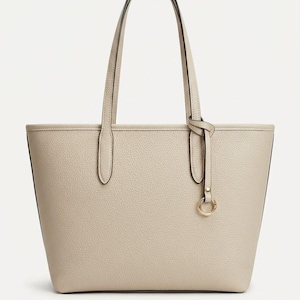 Op de afbeelding: Een beige tote bag met een generfde textuur, voorzien van twee schouderbanden en een decoratieve goudkleurige metalen ringhanger die met een dun riempje aan &eacute;&eacute;n kant is bevestigd. De tas heeft een gestructureerde, trapeziumvormige vorm.