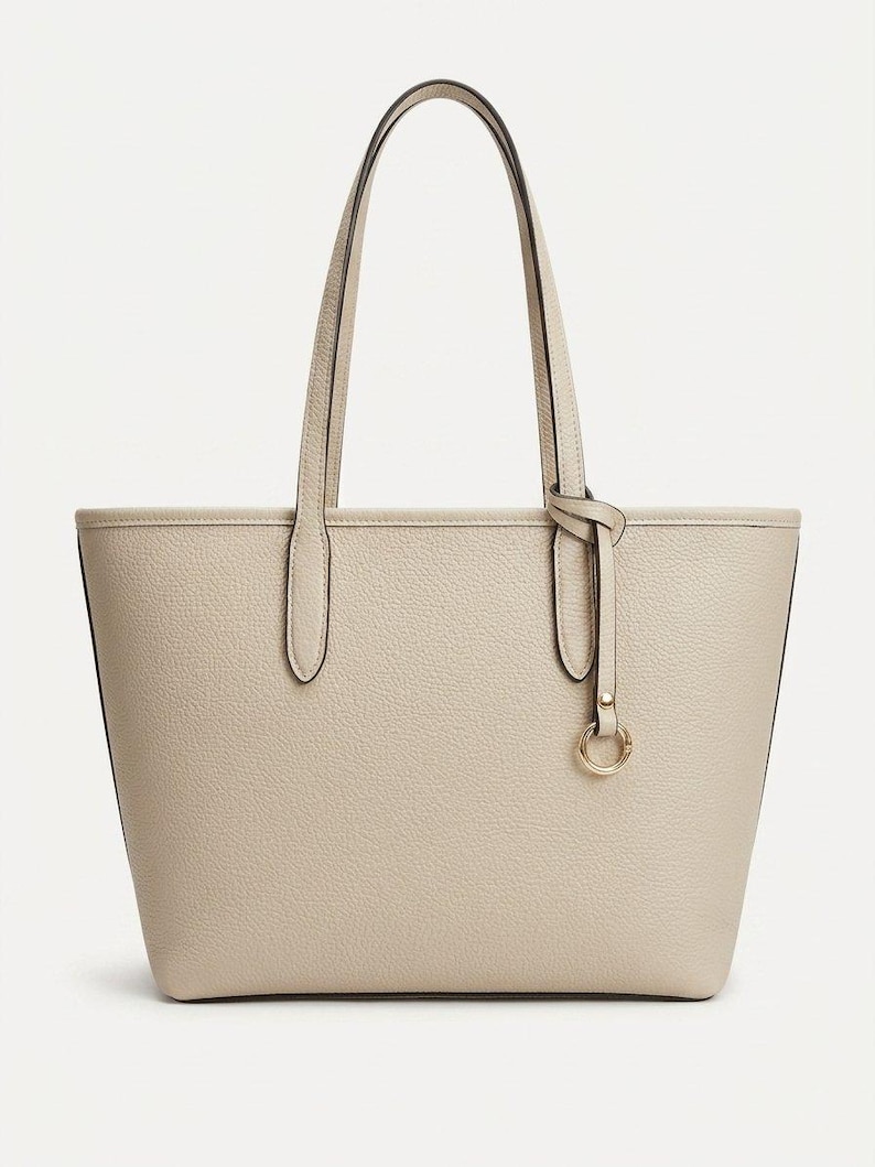 Op de afbeelding: Een beige tote bag met een generfde textuur, voorzien van twee schouderbanden en een decoratieve goudkleurige metalen ringhanger die met een dun riempje aan &eacute;&eacute;n kant is bevestigd. De tas heeft een gestructureerde, trapeziumvormige vorm.