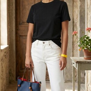 May include: A grey baseball cap, a black short-sleeved t-shirt, white denim jeans, and a navy blue bucket-style handbag with brown leather straps and a bright red interior lining. A yellow wristband is worn on the left wrist.