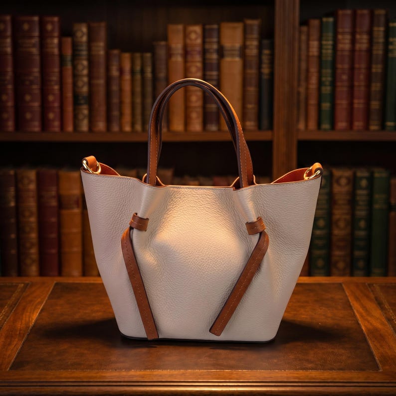 K&ouml;nnte beinhalten: Eine hellbeige Handtasche mit strukturierter Oberfl&auml;che, ausgestattet mit zwei braunen Ledergriffen und zwei dekorativen Knoten-Details aus braunem Leder auf der Vorderseite. Die Tasche hat ein offenes Design mit goldfarbenen Beschl&auml;gen.
