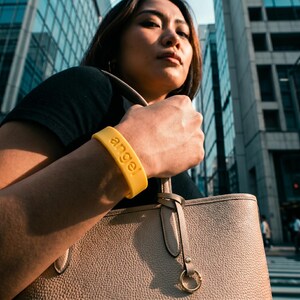 Peut inclure: Un bracelet en silicone jaune vif portant le mot angel en relief en minuscules. Le bracelet est port&eacute; sur l'avant-bras, au-dessus d'un sac fourre-tout en cuir beige textur&eacute; dot&eacute; de finitions dor&eacute;es et d'un d&eacute;tail en forme d'anneau circulaire.