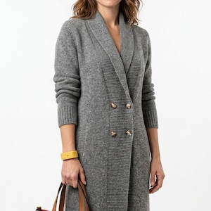 May include: A long grey knit cardigan with a shawl collar and four buttons, paired with a navy blue handbag featuring tan leather handles and a red interior lining. A yellow bracelet is worn on the wrist, completing the ensemble.