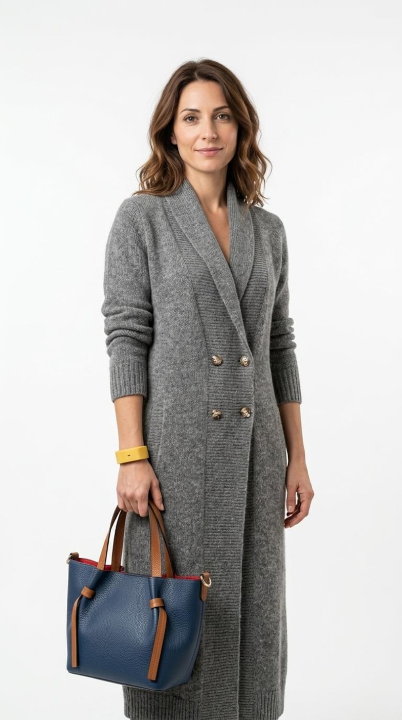 May include: A long grey knit cardigan with a shawl collar and four buttons, paired with a navy blue handbag featuring tan leather handles and a red interior lining. A yellow bracelet is worn on the wrist, completing the ensemble.