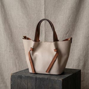 K&ouml;nnte beinhalten: Eine beigefarbene Handtasche aus genarbtem Leder mit zwei dunkelbraunen Ledergriffen und zwei passenden, dekorativen Lederriemen, die an der Vorderseite herabh&auml;ngen. Die Tasche hat ein strukturiertes, offenes Design mit goldfarbenen Beschl&auml;gen.