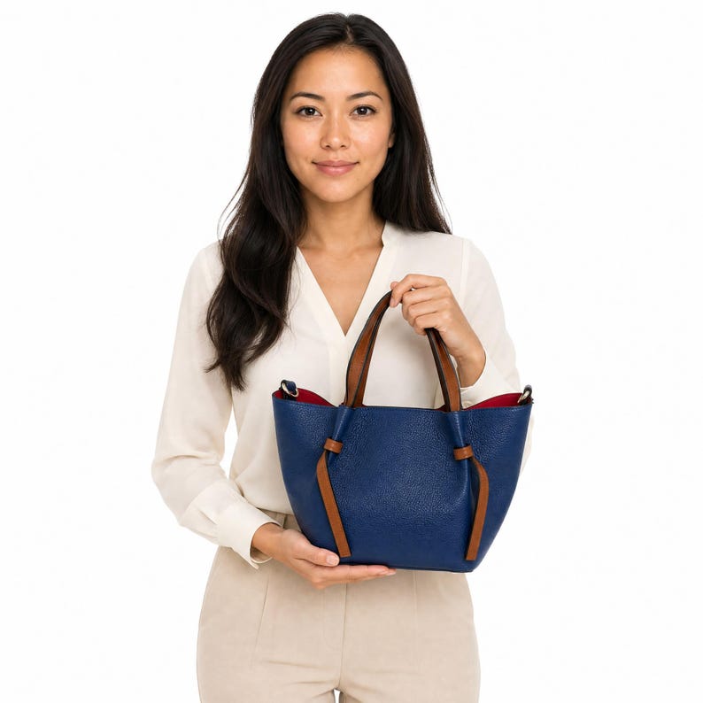K&ouml;nnte beinhalten: Eine blaue Handtasche aus strukturiertem Leder mit zwei braunen Ledergriffen, die an der Basis verknotet sind. Die Tasche hat ein offenes Design mit einem kontrastierenden roten Innenfutter und goldfarbenen Beschl&auml;gen an den Seiten.