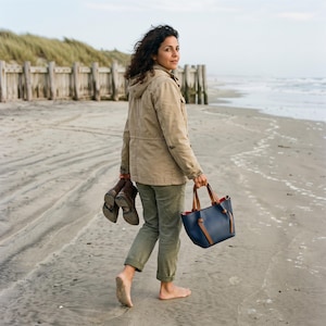K&ouml;nnte beinhalten: Eine beige Kapuzenjacke im Utility-Stil, eine olivgr&uuml;ne Hose und eine marineblaue Ledertasche mit hellbraunen Riemen. Die Tasche hat einen strukturierten Boden und ein leuchtend rotes Innenfutter. Dazu ein Paar dunkelbraune Lederstiefel.