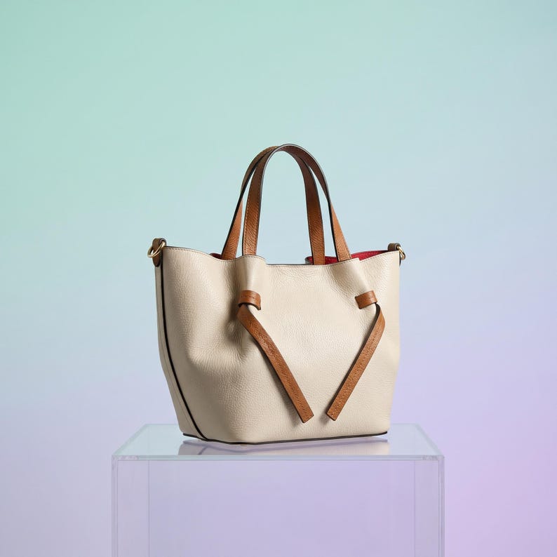 K&ouml;nnte beinhalten: Eine beigefarbene Ledertasche mit zwei braunen Ledergriffen und einem dekorativen, geknoteten braunen Lederriemendetail auf der Vorderseite. Die Tasche hat einen strukturierten Boden, goldfarbene Beschl&auml;ge und ein leuchtend rotes Innenfutter.