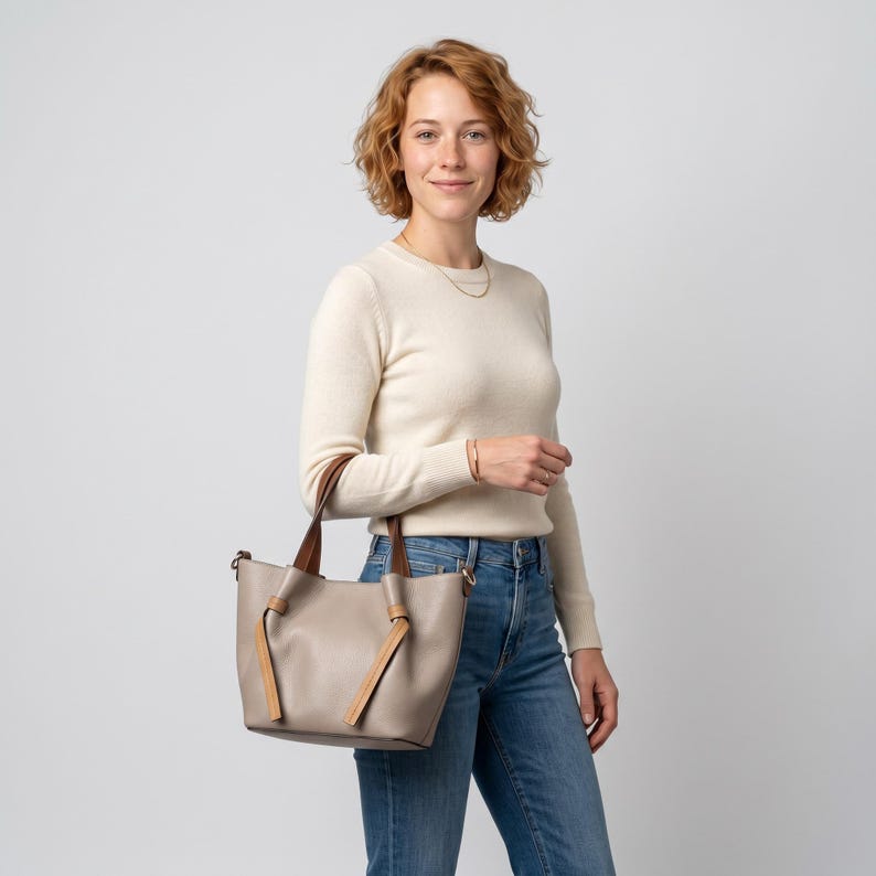 K&ouml;nnte beinhalten: Eine taupefarbene Tragetasche mit zwei braunen Ledergriffen, die an der Vorderseite der Tasche herabh&auml;ngen und ein geknotetes Detail bilden. Die Tasche hat ein strukturiertes Design mit weiter &Ouml;ffnung, glatter Oberfl&auml;che und goldfarbenen Beschl&auml;gen.