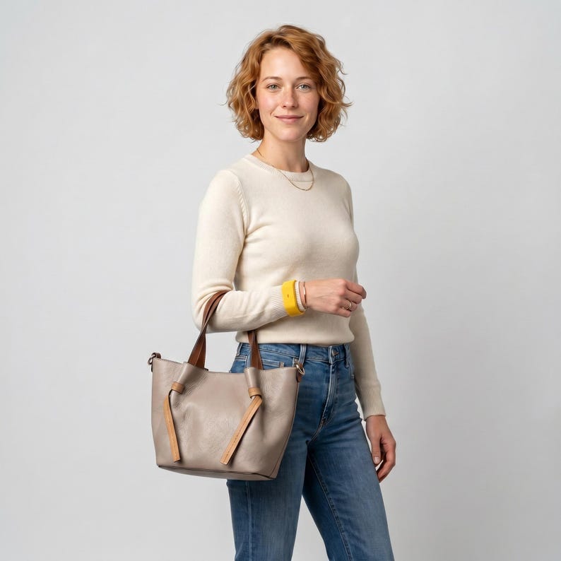 Peut inclure: Un sac cabas de couleur taupe dot&eacute; de deux anses sup&eacute;rieures avec des d&eacute;tails nou&eacute;s &agrave; la base de chaque sangle. Le sac pr&eacute;sente une ouverture large et structur&eacute;e, fabriqu&eacute; dans une mati&egrave;re lisse avec des garnitures couleur cuir beige.