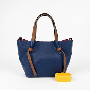 May include: A navy blue leather handbag with a pebbled texture, featuring two brown leather handles and decorative brown leather side ties. The interior lining is bright red. A yellow plastic wristband is positioned in front of the bag.