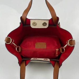 K&ouml;nnte beinhalten: Eine Tragetasche mit hellbraunen Ledergriffen und einem leuchtend roten Innenfutter. Die Tasche verf&uuml;gt &uuml;ber eine mittige Rei&szlig;verschlusstasche, goldfarbene Metallbeschl&auml;ge und zwei cremefarbene Druckknopfverschl&uuml;sse an der oberen &Ouml;ffnung.