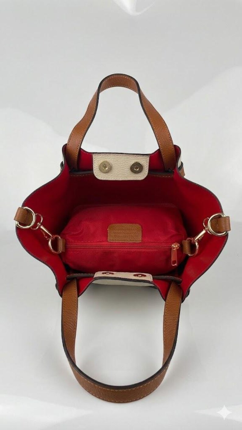 K&ouml;nnte beinhalten: Eine Tragetasche mit hellbraunen Ledergriffen und einem leuchtend roten Innenfutter. Die Tasche verf&uuml;gt &uuml;ber eine mittige Rei&szlig;verschlusstasche, goldfarbene Metallbeschl&auml;ge und zwei cremefarbene Druckknopfverschl&uuml;sse an der oberen &Ouml;ffnung.