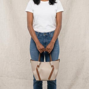 K&ouml;nnte beinhalten: Eine beige Tragetasche mit braunen Ledergriffen und dekorativen, geknoteten braunen Lederakzenten auf der Vorderseite. Die Tasche verf&uuml;gt &uuml;ber einen strukturierten Boden und ein offenes Design, das eine klare, neutrale Farbpalette und eine glatte Oberfl&auml;che zeigt.