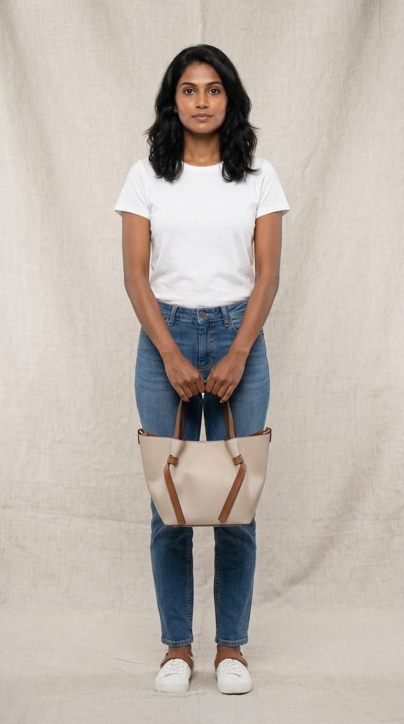 K&ouml;nnte beinhalten: Eine beige Tragetasche mit braunen Ledergriffen und dekorativen, geknoteten braunen Lederakzenten auf der Vorderseite. Die Tasche verf&uuml;gt &uuml;ber einen strukturierten Boden und ein offenes Design, das eine klare, neutrale Farbpalette und eine glatte Oberfl&auml;che zeigt.