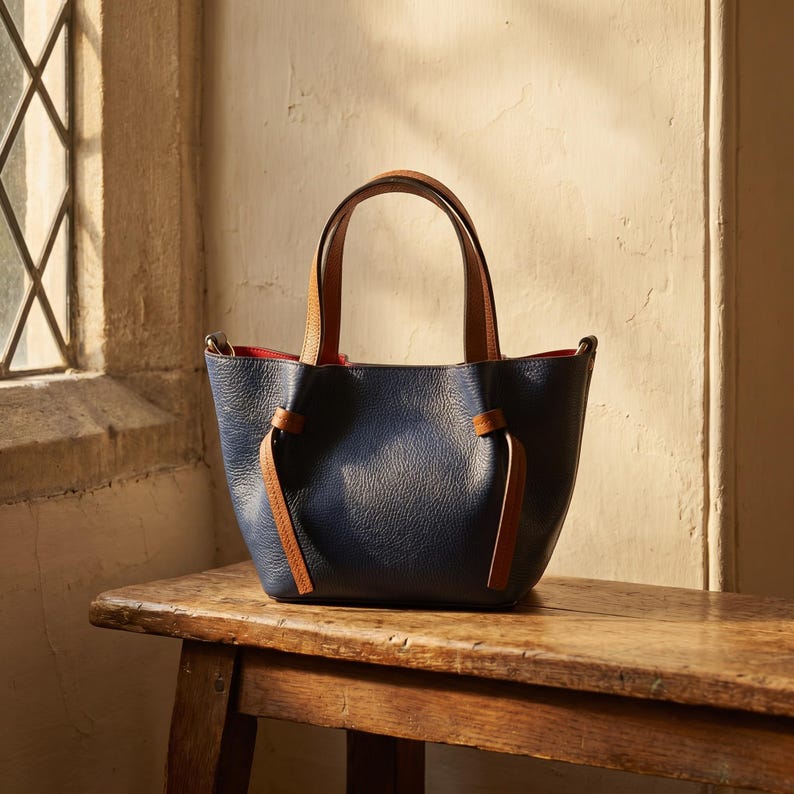May include: A navy blue handbag with a pebbled texture, featuring tan leather handles and matching side knot details. The interior of the bag is lined with a vibrant red material, and the structure includes a wide opening with a curved top edge.