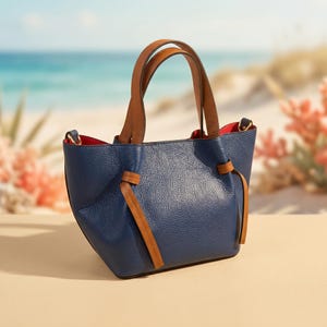 May include: A navy blue pebbled leather handbag featuring two tan leather top handles and a matching tan leather tie detail on the front. The interior of the bag is lined with a vibrant red material, and the exterior includes gold-tone hardware.
