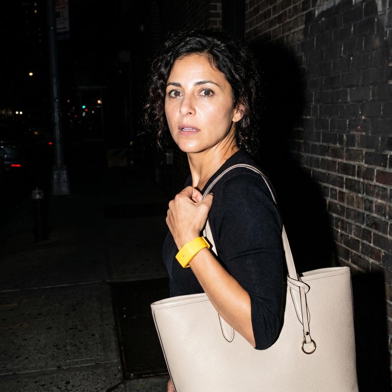 Peut inclure: Un sac cabas beige dot&eacute; de deux bandouli&egrave;res et d'un anneau m&eacute;tallique circulaire, accompagn&eacute; d'un bracelet jaune vif. Le sac pr&eacute;sente une surface textur&eacute;e et est port&eacute; &agrave; l'&eacute;paule, compl&eacute;t&eacute; par un haut noir &agrave; manches longues.
