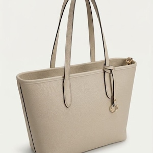 Op de afbeelding: Een beige tote bag met een generfde textuur, voorzien van twee lange schouderbanden en een decoratieve hanger met een goudkleurig ringdetail. De tas heeft een gestructureerd silhouet met strakke lijnen en een eenvoudig open ontwerp.