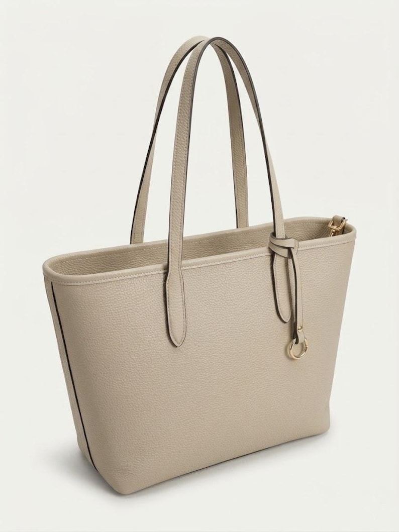 Op de afbeelding: Een beige tote bag met een generfde textuur, voorzien van twee lange schouderbanden en een decoratieve hanger met een goudkleurig ringdetail. De tas heeft een gestructureerd silhouet met strakke lijnen en een eenvoudig open ontwerp.