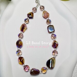 May include: A phone charm strap featuring a series of iridescent, multi-shaped beads in shades of brown, bronze, and purple, interspersed with small clear faceted beads. The text "Lil Bead Story a little bit of story" is printed in the centre.