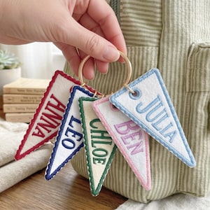 May include: A gold-toned metal ring holding five triangular felt pennant keychains. Each white felt pennant features embroidered names in different colors: red for ANNA, blue for LEO, green for CHLOE, pink for BEN, and light blue for JULIA.