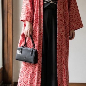 May include: A long red and white patterned kimono-style duster coat worn over a cream-colored high-neck blouse, a wide black waist belt with a cord tie, black wide-leg trousers, and black heeled ankle boots, carrying a small black handbag.