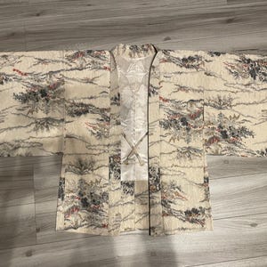 Pode incluir: Uma jaqueta haori de cor creme em tecido texturizado com um padrão de paisagem em tons de cinza, preto e vermelho. A peça possui mangas largas, frente aberta e um forro branco com um padrão floral sutil e dois laços de tecido para fechar.
