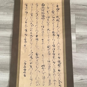 Op de afbeelding: Een verticale hangende rol met traditionele Japanse kalligrafie in zwarte inkt op een lichtbeige papieren achtergrond. De rol is aan de boven- en onderkant omlijst met donkerbruine stoffen randen en heeft een dun koord om op te hangen.