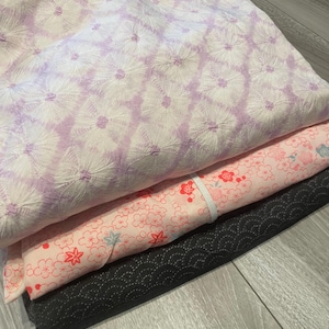 May include: Three folded pieces of fabric stacked together. The top layer is white with a purple tie-dye style sunburst pattern. The middle layer is light pink with a red and blue floral print. The bottom layer is solid black with a textured pattern.