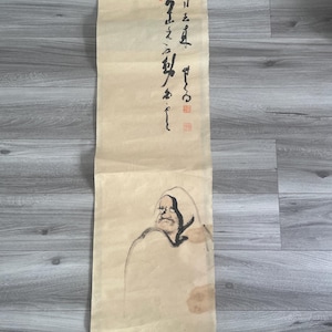 May include: A vertical hanging scroll made of light beige paper featuring black ink brush calligraphy and a minimalist ink drawing of a hooded figure. The artwork includes two red square seals and a wooden roller at the bottom for display.