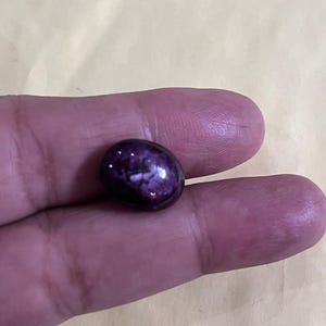 Può includere: Una gemma lucida, di forma ovale, con una tonalità viola intenso e motivi a vortice. La pietra è tenuta tra due dita, mettendo in risalto la sua superficie liscia e riflettente. Il colore della gemma varia dal viola scuro a tonalità più chiare.