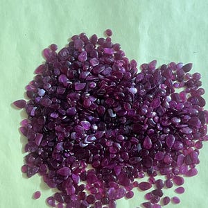 Op de afbeelding: Een stapel gepolijste, diep magenta edelstenen. De stenen variëren in grootte en vorm, met een mix van afgeronde en druppelvormen. De edelstenen liggen verspreid op een lichtgroen oppervlak, wat een contrast creëert.
