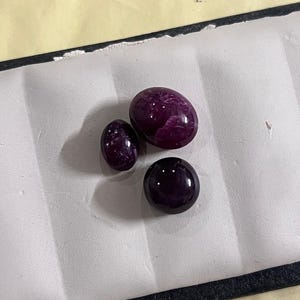 Peut inclure: Trois pierres précieuses polies de tailles variables. Les pierres sont d'un violet profond et riche, avec une surface lisse et réfléchissante. Les pierres précieuses sont exposées sur une surface blanche.