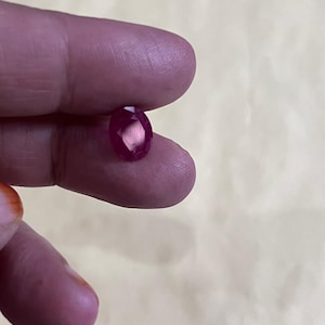 Könnte beinhalten: Ein facettierter, oval geformter Rubin-Edelstein. Der Rubin hat eine tiefe, gesättigte rote Farbe mit einer polierten Oberfläche, die das Licht reflektiert. Der Edelstein wird zwischen den Fingern gehalten, um seine Größe und Form zu zeigen.
