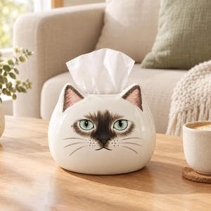 May include: A ceramic tissue box cover shaped like a Siamese cat head with cream-coloured fur, a dark brown face mask, pointed ears with pink inner details, and light green eyes. A white tissue is pulled through the opening at the top of the head.