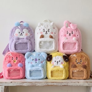 May include: A collection of eight plush backpacks in various colors including lavender, white, pink, blue, yellow, and brown. Each backpack features a soft, fuzzy texture, a clear rectangular front pocket, and a cartoon character face design.